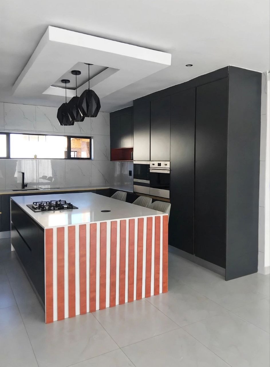 Kitchen with geometric drop ceiling, black faceted pendants, matte black cabinetry, and an island with an orange slatted front and gas cooktop
